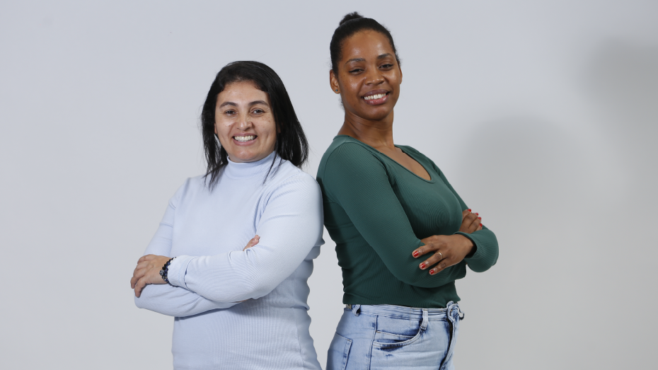 Foto com fundo cinza e duas mulheres de braços cruzados e sorrindo.