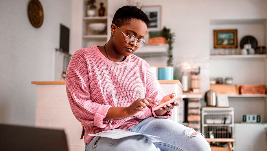 Jovem sentada com o celular na mão e com o computador enquanto analisa informações.