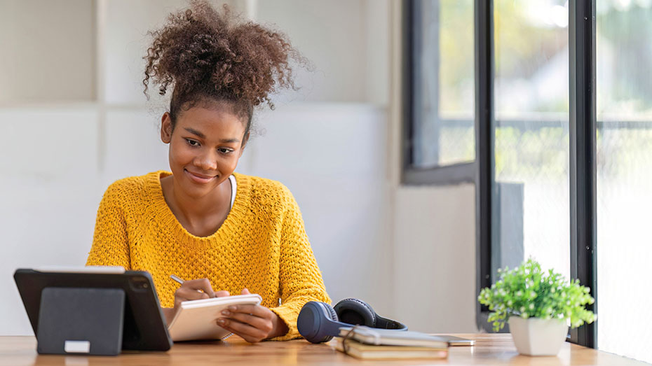 Foto de uma estudante negra que está sorrindo enquanto acessa o Universo CIEE e anota as dicas relacionadas ao mundo do trabalho.