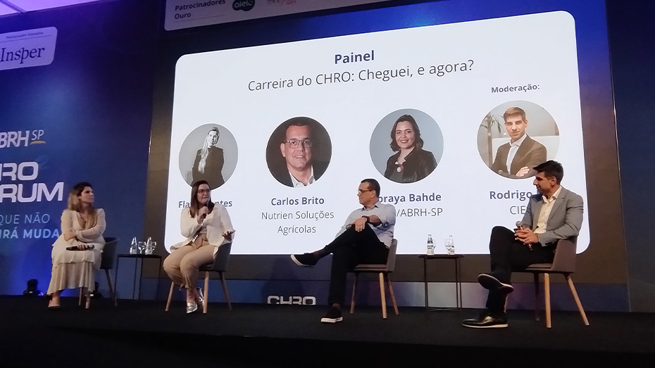 Imagem de um palco com quatro painelistas em telão ao fundo.