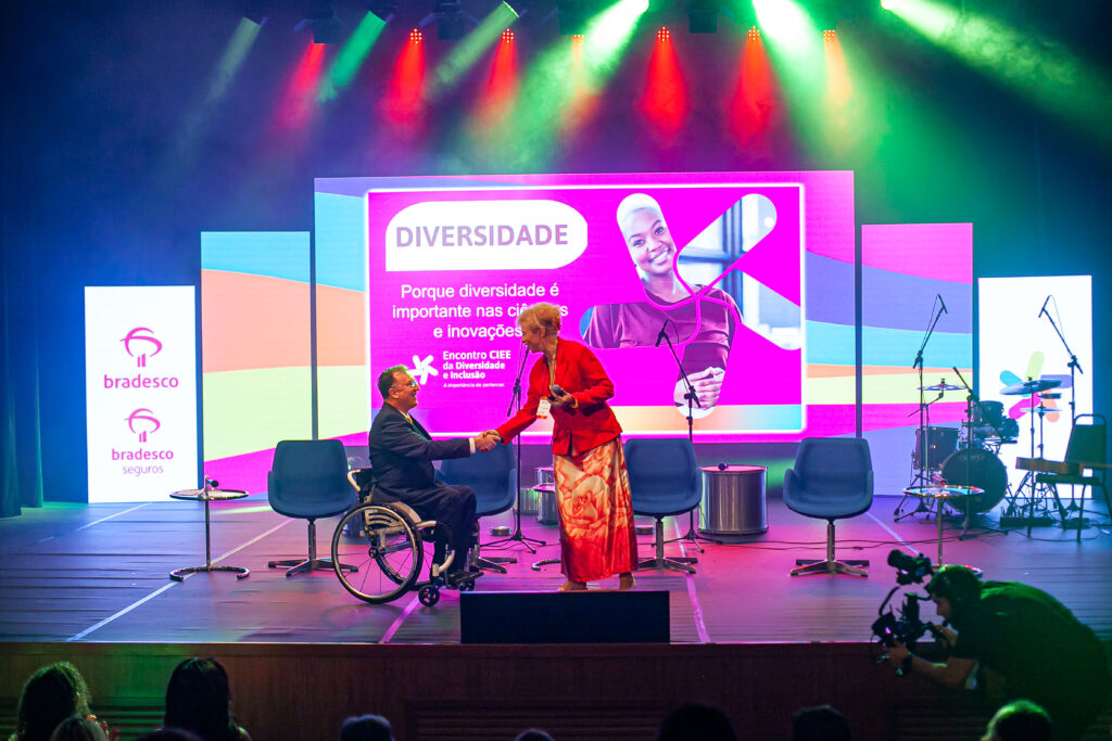 Imagem de palco com um painel ao fundo e duas pessoas ao palco, uma delas um cadeirante.