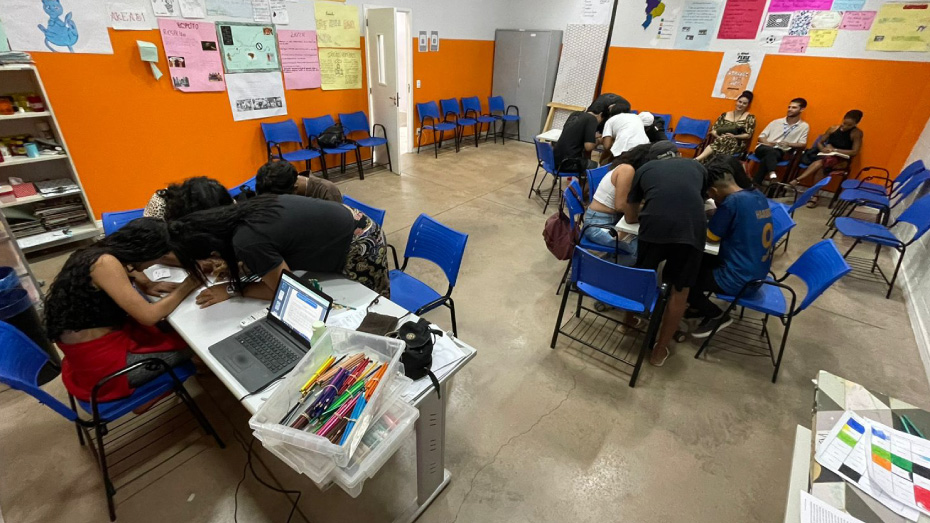 Foto de uma sala com adolescentes divididos em dois grupos realizando uma atividade e ao fundo, três pessoas observando