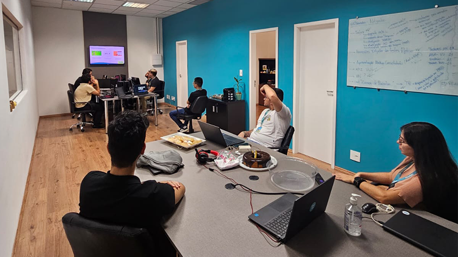 Foto de uma sala retangular com estagiários divididos em duas mesas. Todos estão olhando para o final da sala, para uma imagem apresentada em uma TV.