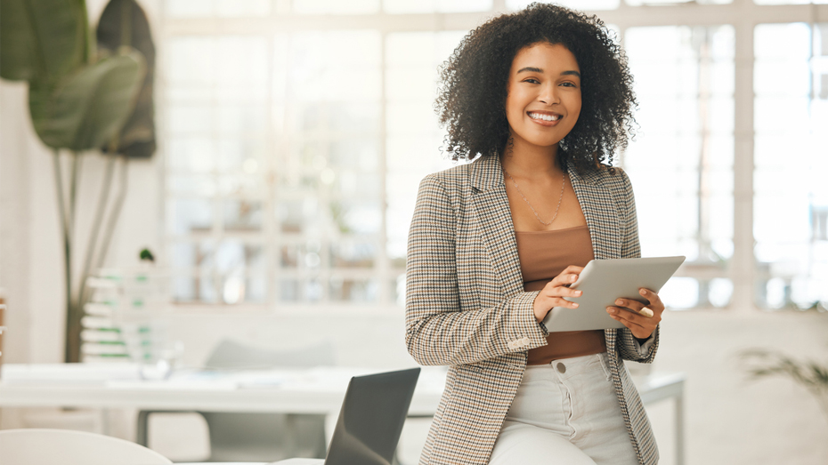 Foto de uma mulher negra sorrindo e segurando um tablet. Ela está em um escritório, usando roupas mais formais.