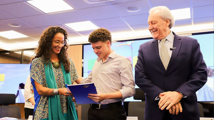 Em uma sala de conferências bem iluminada, Luana Bottini, assessora de Projetos Sociais do CIEE, sorri enquanto entrega um certificado a um jovem de camisa social clara, que também sorri ao recebê-lo. Ao lado deles, o ministro da Justiça e Segurança Pública, Ricardo Lewandowski, de terno escuro e gravata clara com estampa discreta, observa a cena com um sorriso. Ao fundo, há telões com imagens coloridas e outros participantes desfocados.