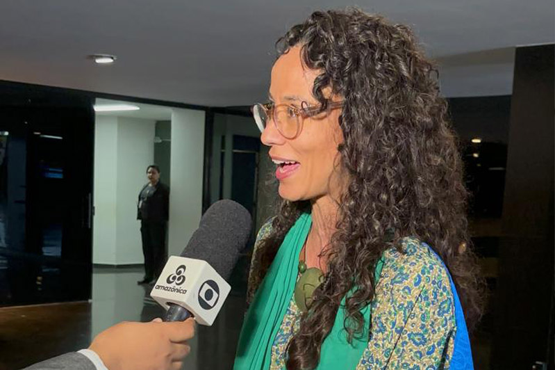 Luana Bottini, assessora de Projetos Sociais do CIEE, dá entrevista a um repórter da TV Globo Amazônica. Ela aparece de perfil, sorrindo e falando, enquanto segura as mãos diante do corpo. Veste um vestido floral azul e verde com uma echarpe verde e um tecido azul sobre os ombros. Usa óculos de armação clara e acessórios grandes, como anéis e braceletes. Ao fundo, o ambiente interno tem iluminação suave e paredes escuras; um segurança aparece desfocado próximo a uma porta de vidro.