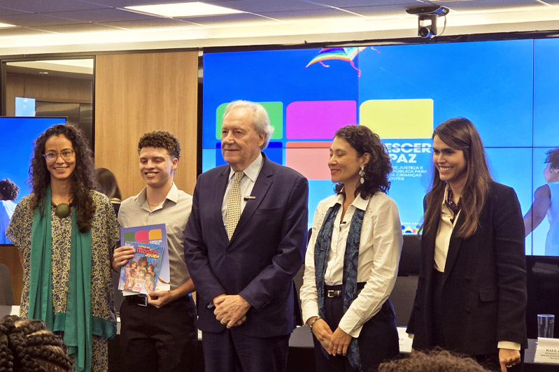 Em um auditório, cinco pessoas estão em pé, lado a lado, sorrindo para a foto. À esquerda, Luana Bottini, assessora de Projetos Sociais do CIEE, usa vestido estampado azul com verde e uma echarpe verde. Ao lado dela, um jovem de camisa clara segura uma cartilha colorida com o título “Manual do Mundo Jovem”. No centro está o ministro da Justiça e Segurança Pública, Ricardo Lewandowski, usando terno azul-marinho e gravata clara. À direita, duas mulheres — uma de camisa branca e lenço azul, a outra de blazer preto — completam o grupo. Ao fundo, telas exibem o logo do programa “Crescer com Paz” e a imagem de uma criança de costas, correndo.